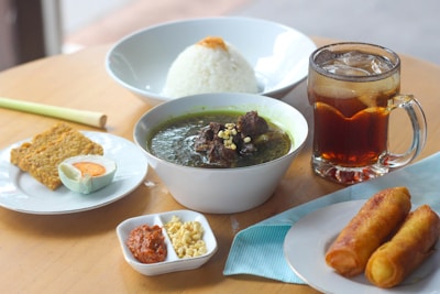A wooden table is set with a variety of Indonesian dishes. A bowl of dark green soup with beef chunks is placed in the center, accompanied by a bowl of white rice in the background. On a small white plate, there are pieces of tempeh and half of a salted egg. A small compartmentalized dish contains chili sambal and chopped nuts. Two spring rolls are placed on a separate plate to the right. A glass mug filled with iced tea, complete with ice cubes, is also present.