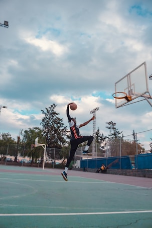A basketball player is captured in mid-air while performing a dunk on an outdoor court. The sky is partly cloudy with patches of blue visible. Surrounding the court are trees and a chain-link fence. Another person sits off to the side of the court, seemingly observing the game.