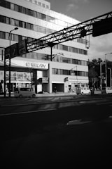 A large multi-story building with numerous windows and a 'CATV' sign is flanked by a busy urban street. Several vehicles, including cars and a bicycle, are visible. The sunlight casts shadows on the building and street, creating a contrast between light and dark areas.
