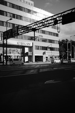 A large multi-story building with numerous windows and a 'CATV' sign is flanked by a busy urban street. Several vehicles, including cars and a bicycle, are visible. The sunlight casts shadows on the building and street, creating a contrast between light and dark areas.