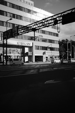 A large multi-story building with numerous windows and a 'CATV' sign is flanked by a busy urban street. Several vehicles, including cars and a bicycle, are visible. The sunlight casts shadows on the building and street, creating a contrast between light and dark areas.