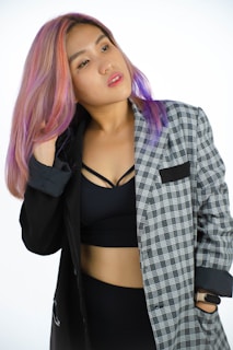 A person with multi-colored hair wearing a black and grey plaid blazer over a black crop top. The individual is adjusting their hair with one hand and looking slightly away, creating a confident and stylish impression.