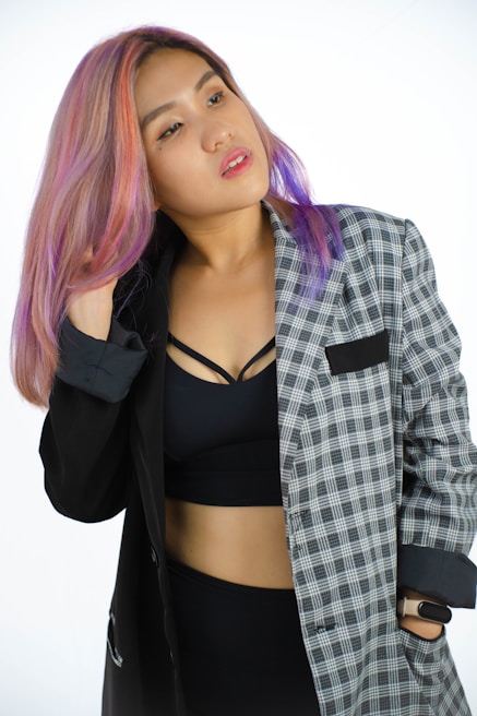 A person with multi-colored hair wearing a black and grey plaid blazer over a black crop top. The individual is adjusting their hair with one hand and looking slightly away, creating a confident and stylish impression.