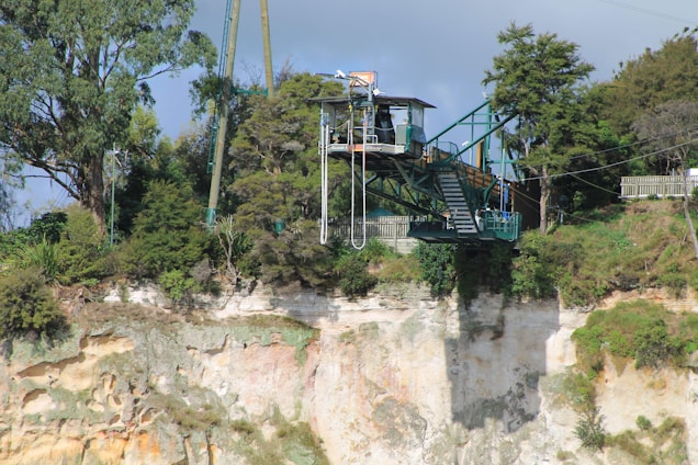 A bungee jumping platform is perched on the edge of a cliff surrounded by lush green trees and vegetation. The structure has stairs leading up to it and is well-integrated into the natural environment. The cliff face is visible with varying shades of rock, and the sky is partly cloudy.
