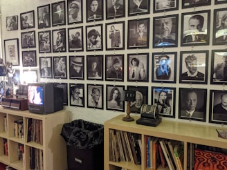 A wall is adorned with a grid of black and white photographs, each featuring different individuals in various poses and expressions. Below the photographs, a shelf holds books, records, a small vintage television, and an old-fashioned cash register. A projector is situated on the left, and a trash bin is visible beneath the shelf.