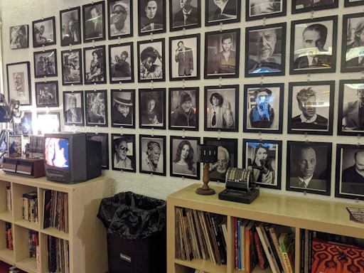 A wall is adorned with a grid of black and white photographs, each featuring different individuals in various poses and expressions. Below the photographs, a shelf holds books, records, a small vintage television, and an old-fashioned cash register. A projector is situated on the left, and a trash bin is visible beneath the shelf.
