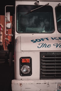 The front end of a vintage ice cream truck with visible headlight and black grille. The truck is painted white with blue text partially reading 'SOFT ICE CREAM.' Reflections and shadows indicate it is parked in an urban environment.