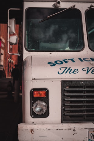 The front end of a vintage ice cream truck with visible headlight and black grille. The truck is painted white with blue text partially reading 'SOFT ICE CREAM.' Reflections and shadows indicate it is parked in an urban environment.