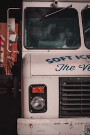 The front end of a vintage ice cream truck with visible headlight and black grille. The truck is painted white with blue text partially reading 'SOFT ICE CREAM.' Reflections and shadows indicate it is parked in an urban environment.