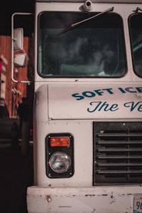 The front end of a vintage ice cream truck with visible headlight and black grille. The truck is painted white with blue text partially reading 'SOFT ICE CREAM.' Reflections and shadows indicate it is parked in an urban environment.