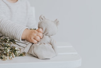 A child's hand gently holds a soft, fabric plush toy, resembling a bear. Beside the toy, there are small, delicate white flowers on slender stems. The background and the child's clothing are in a soft, neutral tone, creating a serene and peaceful atmosphere.