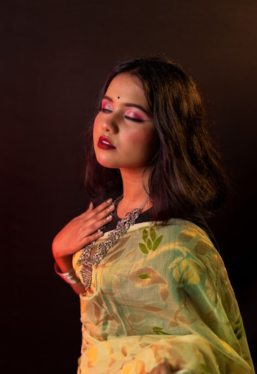 A woman with closed eyes appears serene, wearing traditional attire with intricate jewelry. Her makeup features bold pink eyeshadow and deep red lipstick. The background is dark, enhancing the warm lighting that casts a gentle glow on her skin.