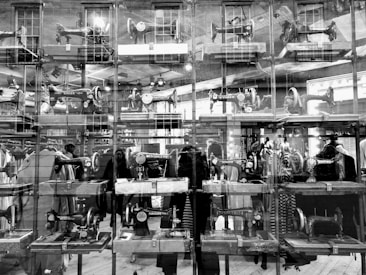A large display case contains multiple vintage sewing machines, each placed on wooden platforms. The glass creates reflections of the surrounding interior, including mannequins and people, giving a layered visual effect. The sewing machines have distinctive brands like Singer and others visible.