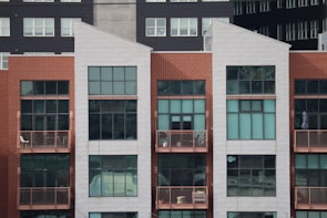 A modern, multi-story apartment building with a combination of red brick and white paneling. The front facade features large rectangular windows and small balconies with railings. Some balconies have furniture and decor items, including chairs and potted plants.