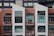 A modern, multi-story apartment building with a combination of red brick and white paneling. The front facade features large rectangular windows and small balconies with railings. Some balconies have furniture and decor items, including chairs and potted plants.