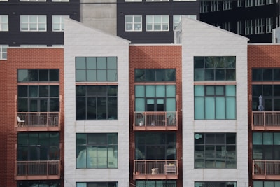 A modern, multi-story apartment building with a combination of red brick and white paneling. The front facade features large rectangular windows and small balconies with railings. Some balconies have furniture and decor items, including chairs and potted plants.