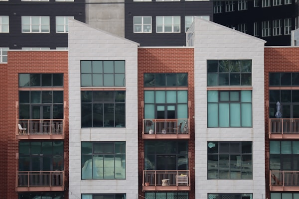 A modern, multi-story apartment building with a combination of red brick and white paneling. The front facade features large rectangular windows and small balconies with railings. Some balconies have furniture and decor items, including chairs and potted plants.