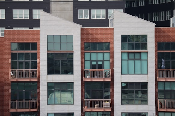 A modern, multi-story apartment building with a combination of red brick and white paneling. The front facade features large rectangular windows and small balconies with railings. Some balconies have furniture and decor items, including chairs and potted plants.