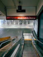 A tiled airport corridor leads to an escalator descending into the space below. Above the escalator is a large red sign displaying 'Welcome to Philadelphia' with a skyline graphic. The lighting is soft, giving a calm atmosphere to the area.