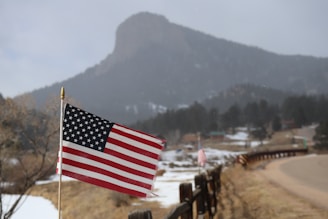 An American flag on a pole prominently displayed in the foreground, with a rural landscape characterized by a snow-dusted field and a mountain in the background. A country road winds alongside a series of fences, giving a sense of vastness and openness.