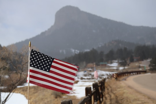An American flag on a pole prominently displayed in the foreground, with a rural landscape characterized by a snow-dusted field and a mountain in the background. A country road winds alongside a series of fences, giving a sense of vastness and openness.