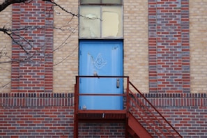 A blue door with a Masonic symbol is set in a multi-colored brick wall. A metal staircase leads up to the door, and a bare tree branch extends into the frame from the left side.