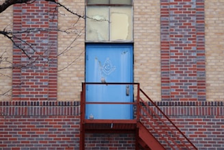 A blue door with a Masonic symbol is set in a multi-colored brick wall. A metal staircase leads up to the door, and a bare tree branch extends into the frame from the left side.