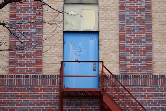 A blue door with a Masonic symbol is set in a multi-colored brick wall. A metal staircase leads up to the door, and a bare tree branch extends into the frame from the left side.