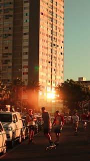 A group of people riding skateboards on a street lined with parked cars. The scene is set in an urban environment with a tall building reflecting the warm glow of the setting sun. Palm trees and other greenery add a touch of nature to the surroundings.