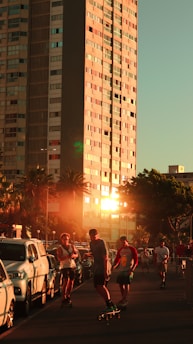 A group of people riding skateboards on a street lined with parked cars. The scene is set in an urban environment with a tall building reflecting the warm glow of the setting sun. Palm trees and other greenery add a touch of nature to the surroundings.