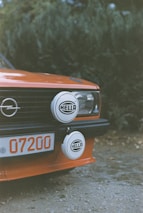 A close-up of the front section of an orange vintage car featuring a pair of round auxiliary lights with the brand name 'HELLA' mounted on the grille. The car's license plate shows the number '07200', and there is a prominent Opel logo on the grille. The background consists of out-of-focus greenery.