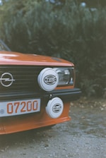 A close-up of the front section of an orange vintage car featuring a pair of round auxiliary lights with the brand name 'HELLA' mounted on the grille. The car's license plate shows the number '07200', and there is a prominent Opel logo on the grille. The background consists of out-of-focus greenery.