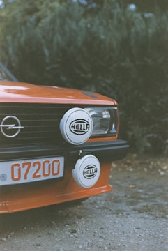 A close-up of the front section of an orange vintage car featuring a pair of round auxiliary lights with the brand name 'HELLA' mounted on the grille. The car's license plate shows the number '07200', and there is a prominent Opel logo on the grille. The background consists of out-of-focus greenery.