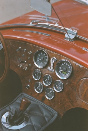 A vintage car dashboard with wooden paneling and chrome-rimmed gauges. The interior also features a gear shift with a wooden handle and classic design elements such as a leather-covered dashboard. The dials include speedometer, fuel gauge, and other measuring instruments, with a distinctive snake emblem visible.