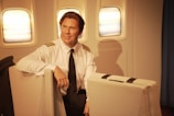 A pilot sits in an airplane cabin, dressed in a white uniform shirt with black shoulder stripes and a black tie. The interior includes three windows, offering a view of the sky with a subtle warm glow.