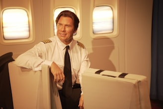 A pilot sits in an airplane cabin, dressed in a white uniform shirt with black shoulder stripes and a black tie. The interior includes three windows, offering a view of the sky with a subtle warm glow.