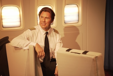 A pilot sits in an airplane cabin, dressed in a white uniform shirt with black shoulder stripes and a black tie. The interior includes three windows, offering a view of the sky with a subtle warm glow.