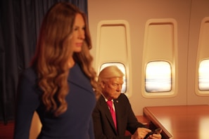 A wax figure of a man is sitting inside an aircraft cabin, looking at a mobile device. The cabin has three windows through which a sunset can be seen. In the foreground, there is a wax figure of a woman in a blue dress with long, wavy hair.
