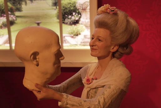A woman with elaborately styled gray hair adorned with pink flowers holds a bald, sculpted head. She wears a light-colored outfit with a shawl, and a smile graces her face. In the background, a window reveals a garden scene with lush greenery.
