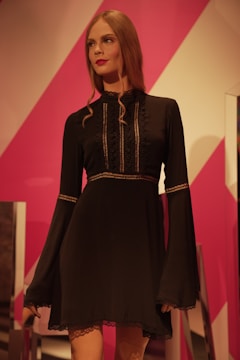 A mannequin dressed in a black, long-sleeved dress with intricate lace detailing on the front and sleeves. The mannequin has long, wavy brown hair and stands against a vibrant pink and white geometric background.
