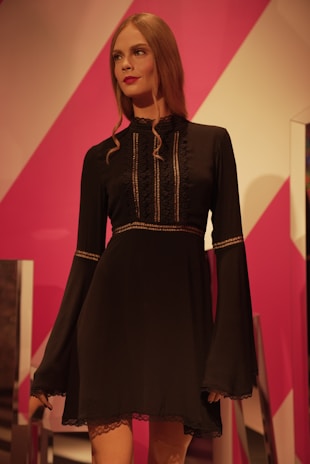 A mannequin dressed in a black, long-sleeved dress with intricate lace detailing on the front and sleeves. The mannequin has long, wavy brown hair and stands against a vibrant pink and white geometric background.