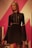 A mannequin dressed in a black, long-sleeved dress with intricate lace detailing on the front and sleeves. The mannequin has long, wavy brown hair and stands against a vibrant pink and white geometric background.