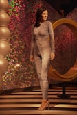 A mannequin wearing a form-fitting, textured bodysuit stands in a stylish setting with vibrant, multicolored sequined walls. The floor features black and white stripes, and there are large spherical decorative elements to the left. A large, golden abstract ring structure is behind the mannequin.
