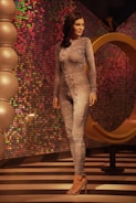 A mannequin wearing a form-fitting, textured bodysuit stands in a stylish setting with vibrant, multicolored sequined walls. The floor features black and white stripes, and there are large spherical decorative elements to the left. A large, golden abstract ring structure is behind the mannequin.