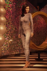 A mannequin wearing a form-fitting, textured bodysuit stands in a stylish setting with vibrant, multicolored sequined walls. The floor features black and white stripes, and there are large spherical decorative elements to the left. A large, golden abstract ring structure is behind the mannequin.