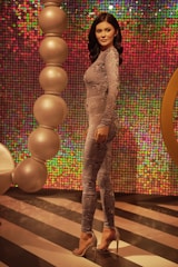 A woman in a patterned grey jumpsuit poses confidently against a vibrant, multicolored sequin wall. She stands on a floor with bold black and white stripes, surrounded by large, spherical decorative elements. Her dark hair is styled in loose waves, and she wears high-heeled shoes.