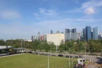 A cityscape with high-rise buildings in the background under a clear blue sky. In the foreground, there is a green lawn bordered by a road with several trees lined up. A small building with a drive-thru setup and a few parked motorcycles is seen on the right side.