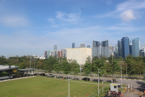 A cityscape with high-rise buildings in the background under a clear blue sky. In the foreground, there is a green lawn bordered by a road with several trees lined up. A small building with a drive-thru setup and a few parked motorcycles is seen on the right side.
