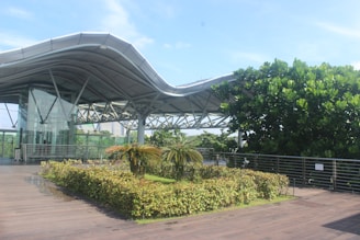 A modern architectural structure with a wavy, metallic roof and a glass-walled elevator is visible. In the foreground, there's a neatly trimmed hedge and a small palm tree surrounded by greenery. The sky is a bright blue, and various trees add to the lush, natural setting.