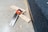 On a speckled concrete floor, a handsaw with an orange and black handle is placed on three pieces of light-colored wooden plank alongside a black carpeted edge.